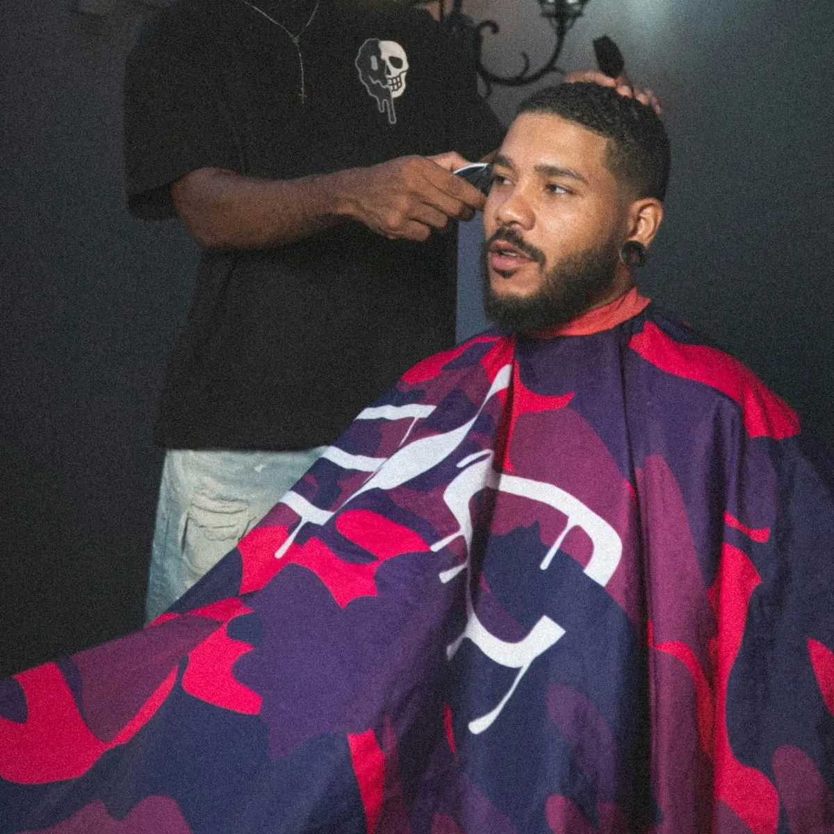 A barber using clippers on a client seated in the chair under a patterned cape
