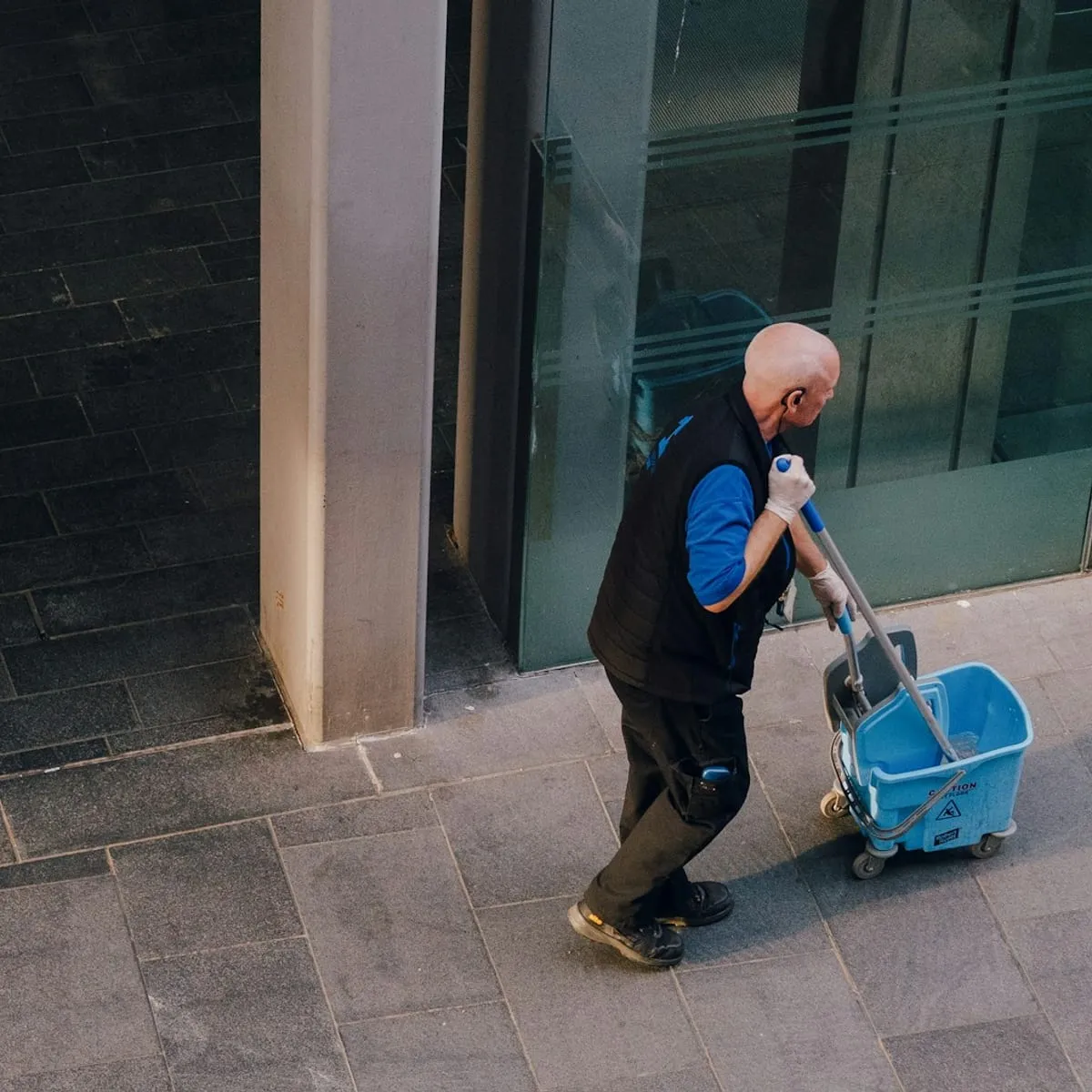 A cleaner pushing a mop and bucket along the outside of a glass office building